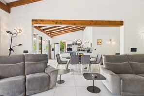 Ein modernes Wohnzimmer mit grauen Sofas, einem Esstisch mit Stühlen, einer Küche im Hintergrund und einem Holzbalken an der Decke.