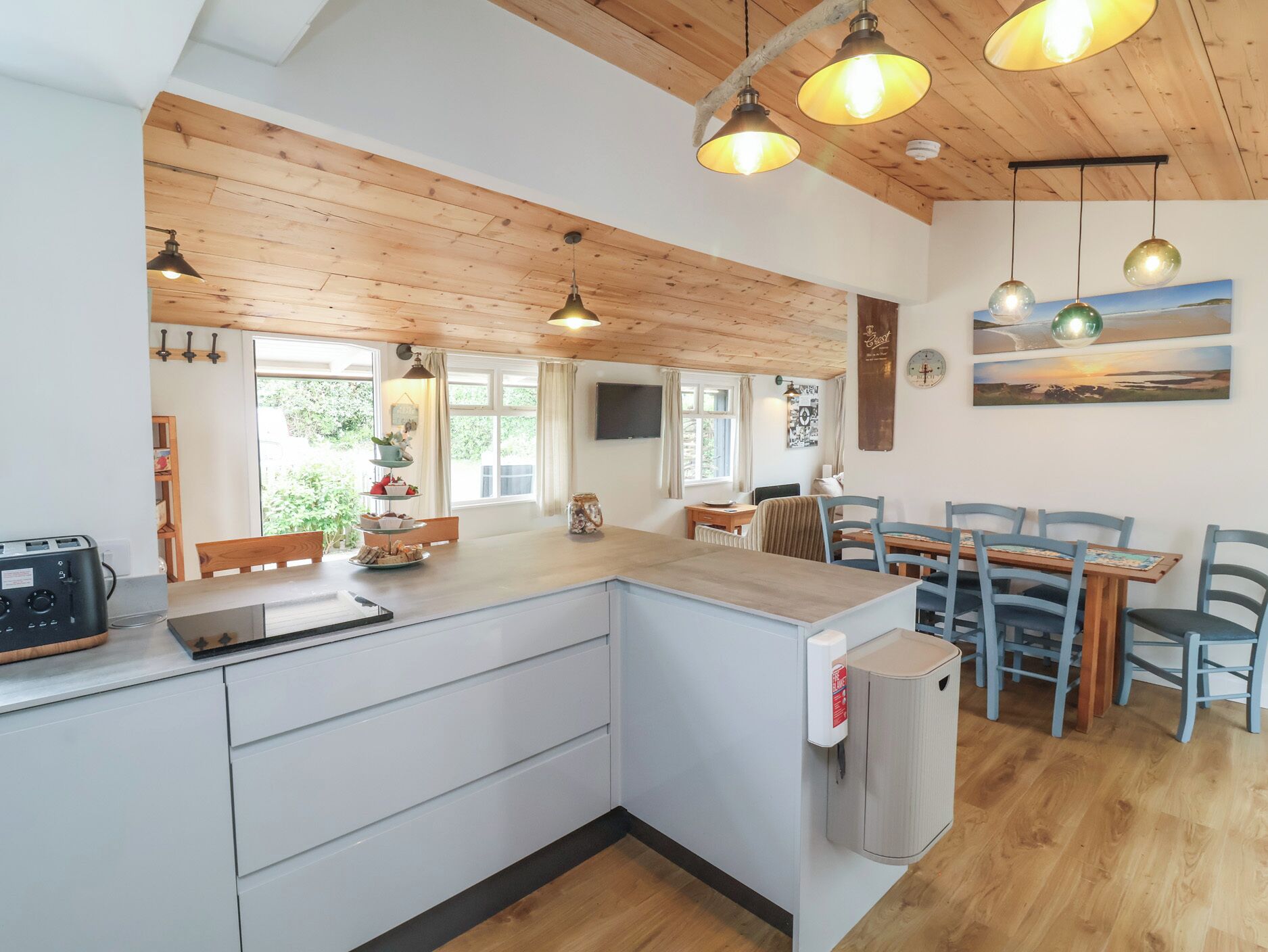 A modern kitchen with wooden beams, pendant lights, a central island, and a dining area with blue chairs.