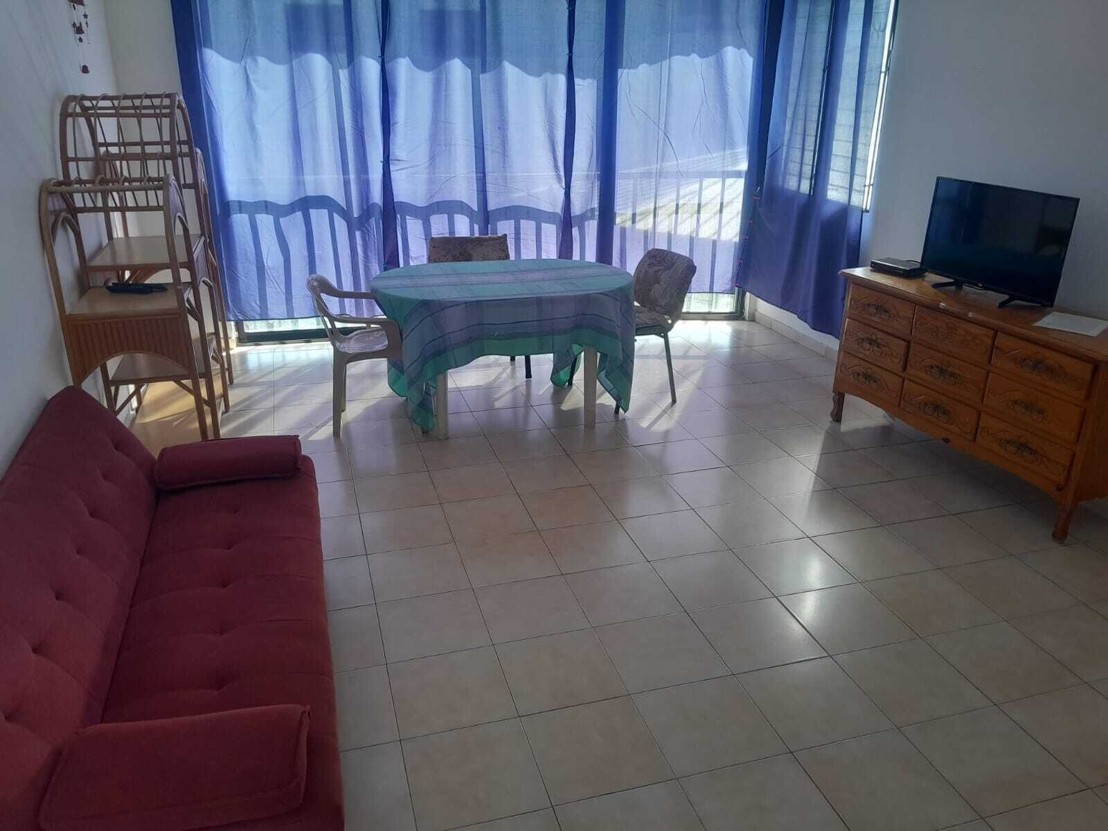 A living room with a red sofa, a dining table with chairs, a wooden cabinet with a TV, and a window with blue curtains.