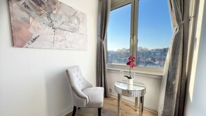A room with a view of a cityscape through a large window, a tufted armchair, a small round table with a vase of flowers, and a large abstract painting on the wall. - Luxury Penthouse, next to Harrods (Londra)