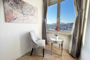 A room with a view of a cityscape through a large window, a tufted armchair, a small round table with a vase of flowers, and a large abstract painting on the wall. - Luxury Penthouse, next to Harrods (Londra)