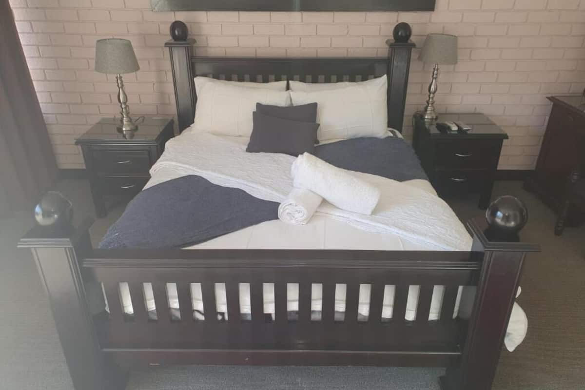 A neatly made bed with a dark wooden frame, white bedding, and a rolled towel. There are two bedside tables with lamps, a dark wooden nightstand, and a brick wall in the background.
