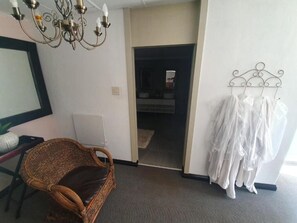 A room with a wicker chair, a white wall, a chandelier, and a clothing rack. - Leach Lodge (Kuruman)