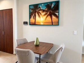 A dining area with a wooden table, two chairs, a vase, a painting of a sunset with palm trees, a TV, and a wardrobe.