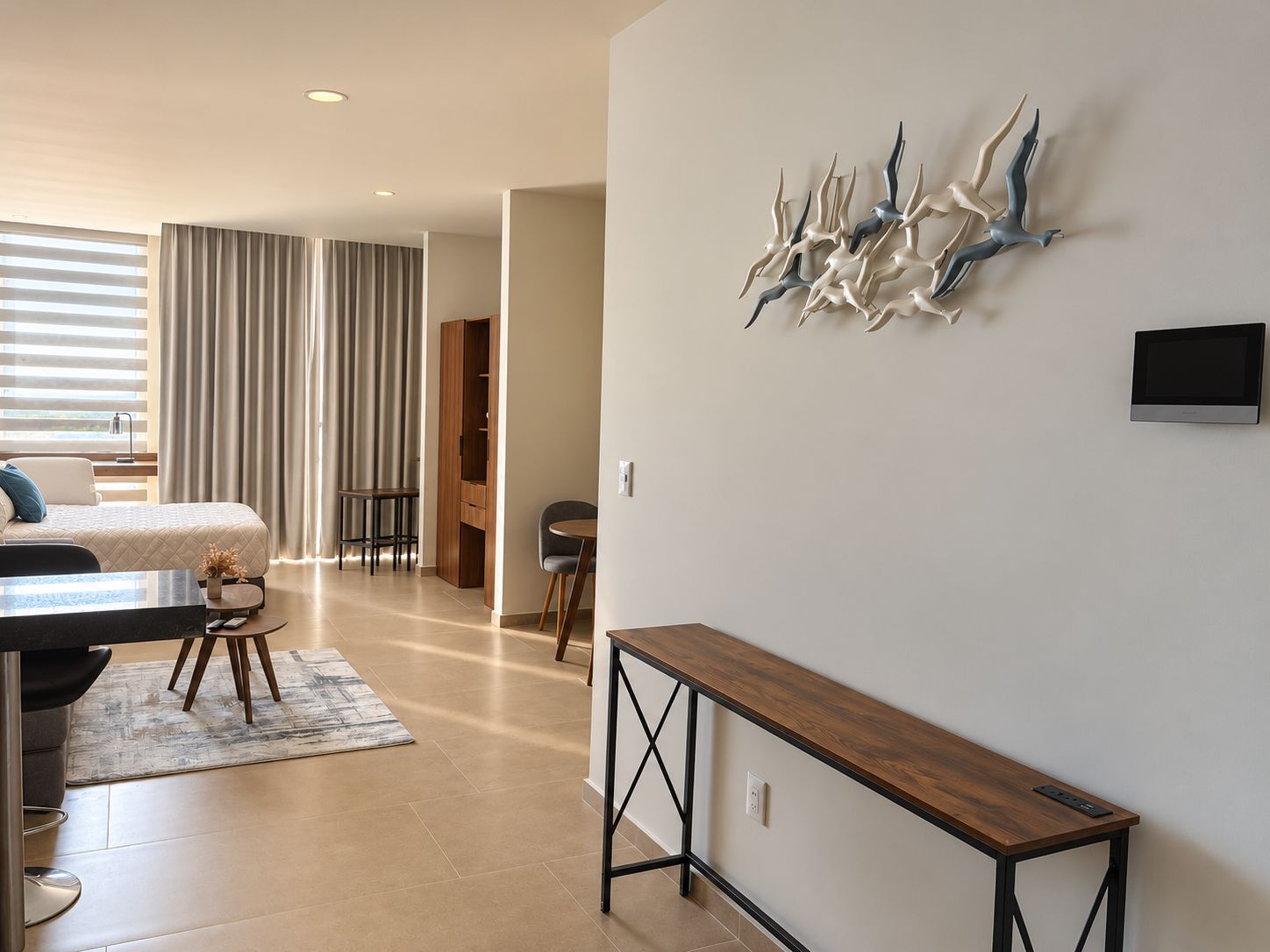 Quarto de hotel moderno com cama, escrivaninha, cadeira, mesa pequena e um objeto decorativo na parede.