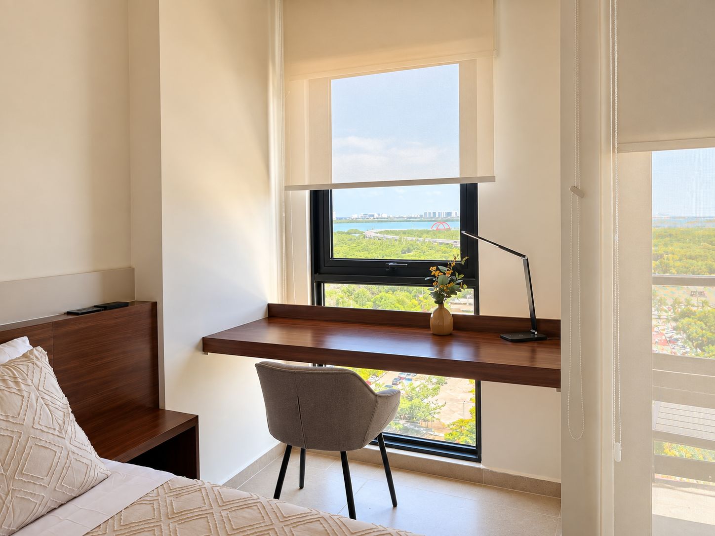 Quarto com cama, escrivaninha com cadeira, janela com vista para vegetação e uma ponte, e mesa de cabeceira.