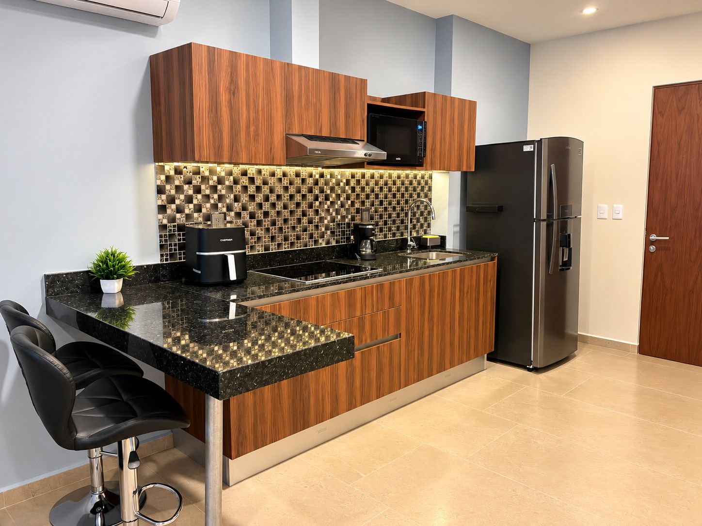 Una cocina moderna con armarios de madera, encimera negra, microondas integrado, cafetera y refrigerador de acero inoxidable.
