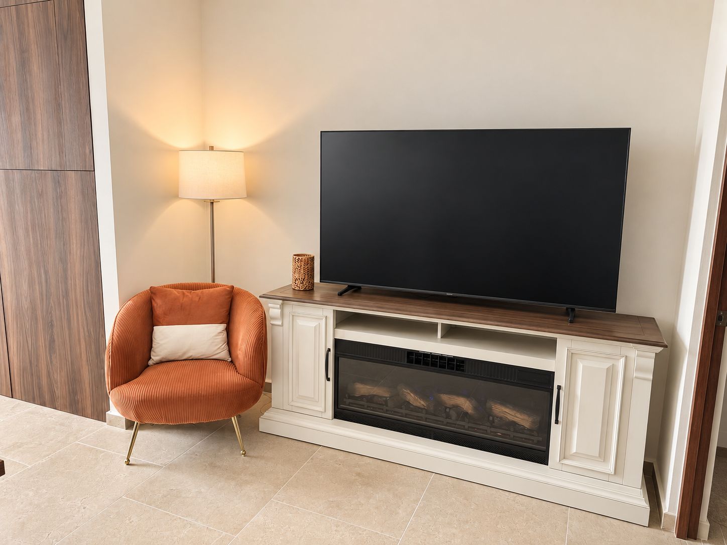 Una sala de estar con un televisor de pantalla plana instalado en un mueble de madera, una lámpara de pie y un sillón con cojines.