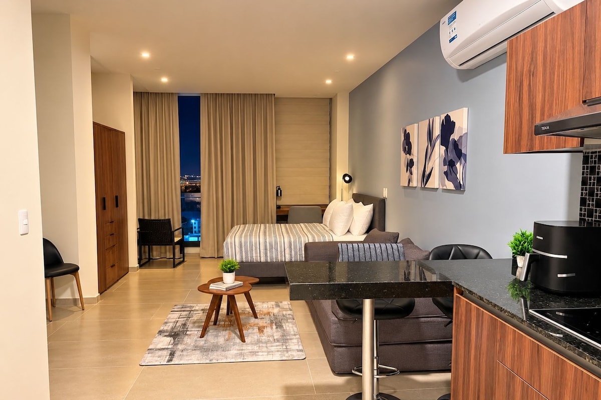 A modern apartment with a kitchenette, a dining area, a living room with a sofa and a bed, and a view of the city at night.