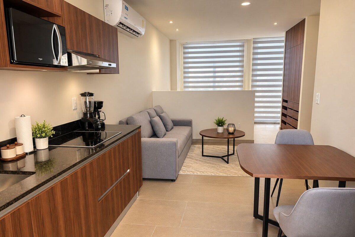 A modern kitchen with a built-in microwave, a coffee maker, and a blender on the counter. There is a grey sofa and a small round table with a plant and candle. A wooden dining table with chairs is also visible.