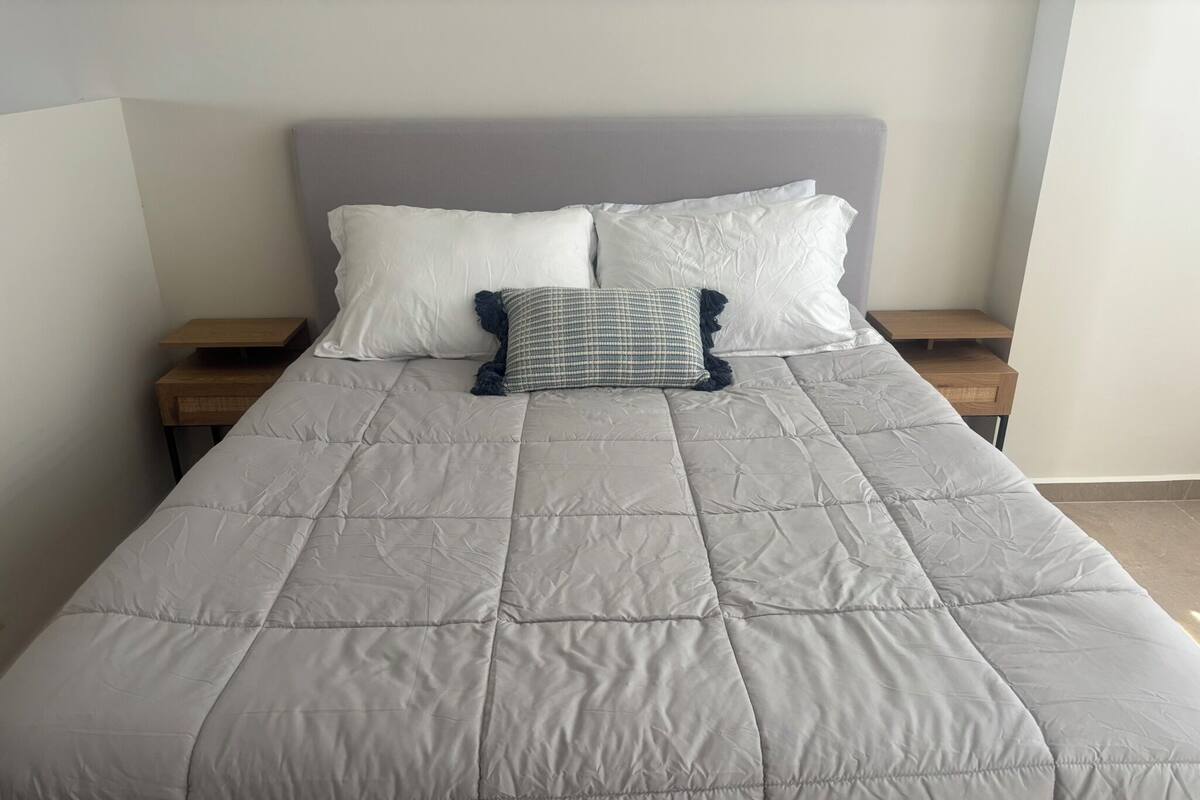 A bed with a quilted grey duvet, two white pillows, and a checkered pillow on a wooden nightstand.