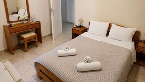 A bedroom with a bed, a wooden dresser, a mirror, a bedside table, and a door to a bathroom. - Hotel Elena -Sunny Seaside Balcony Suite in Skyros (Skyros)