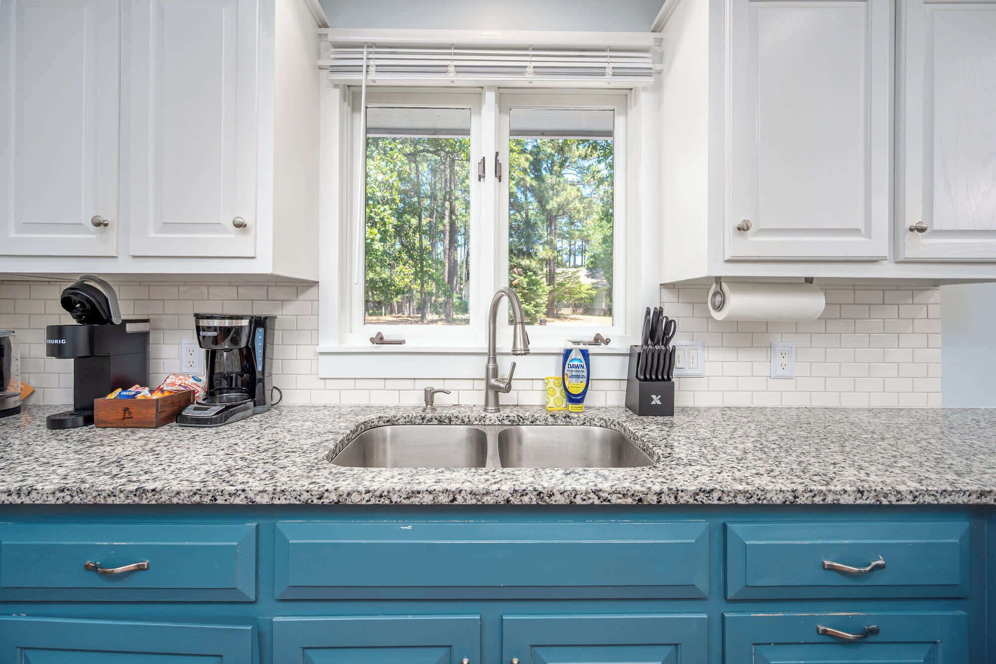 Una cocina con armarios blancos, encimera de granito, pileta de acero inoxidable y ventana con vista a árboles.