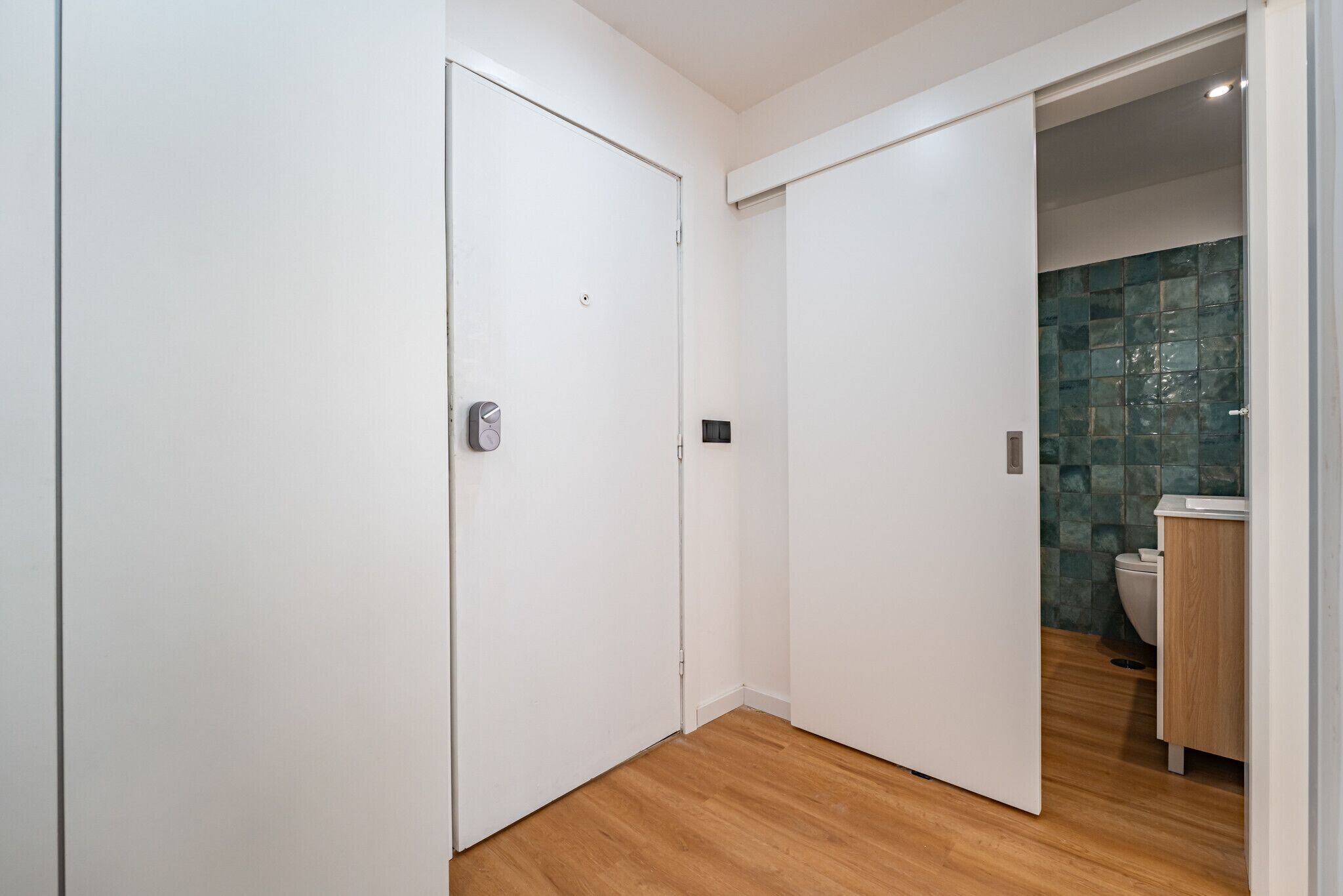 A hallway with a wooden floor, white walls, and a bathroom with a green-tiled wall and a white toilet.