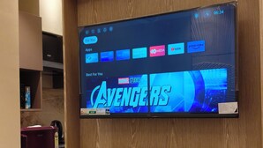 A flat-screen TV mounted on a wooden wall with a 'For You' screen displaying 'AVENGERS' and various app icons.