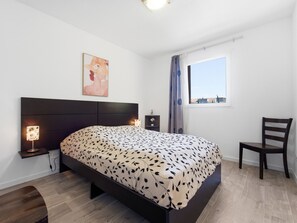 Ein Schlafzimmer mit Bett, Stuhl, Nachttisch, Lampe, Fenster und einem Bild an der Wand.