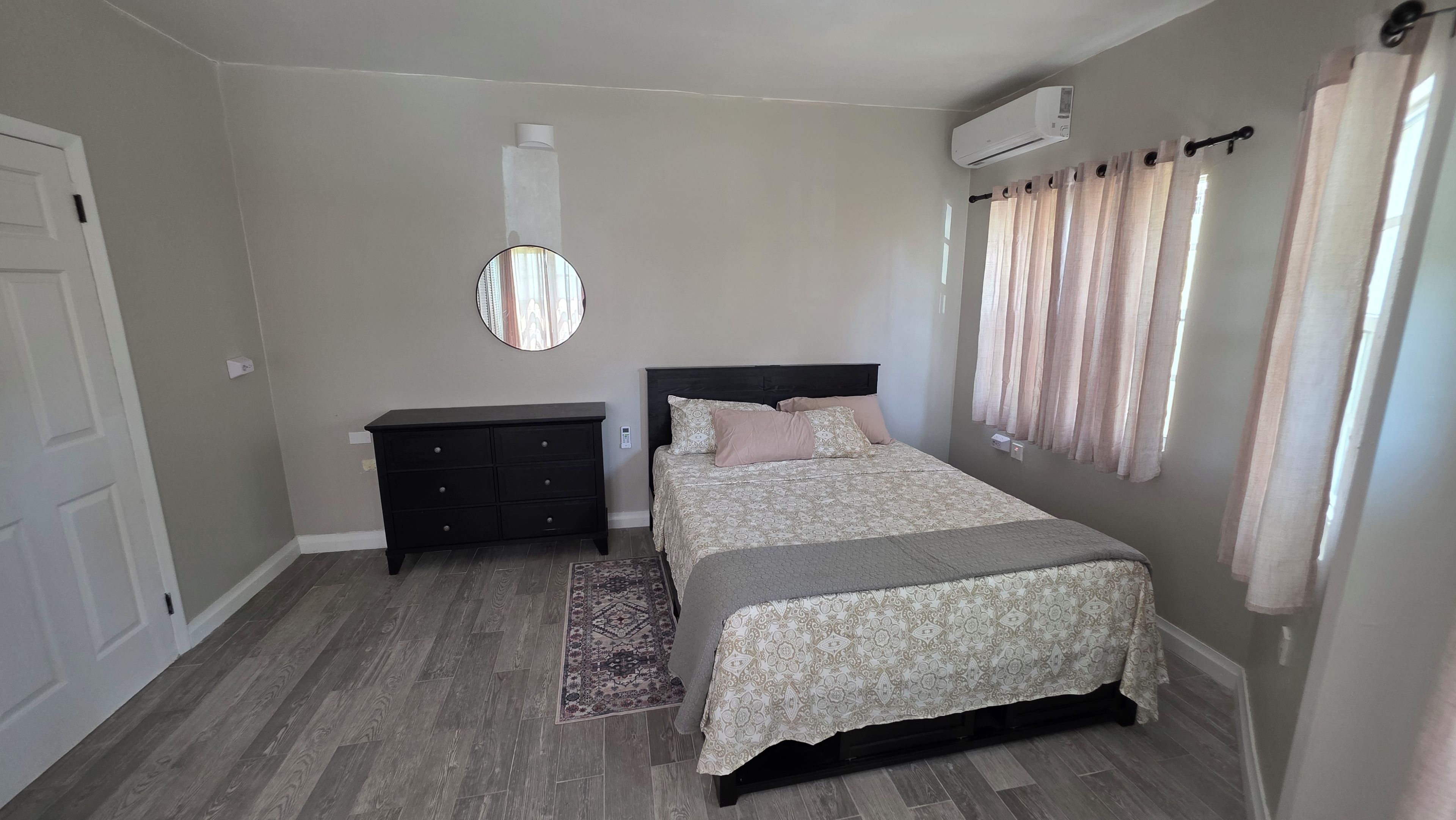 Une chambre à coucher comprenant un lit, une commode, un miroir, un appareil de climatisation et une fenêtre avec des rideaux.