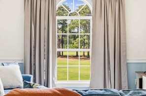 Ein Schlafzimmer mit großem Fenster, Vorhängen, einem Bett mit Kissen und Blick auf eine Rasenfläche und Bäume.