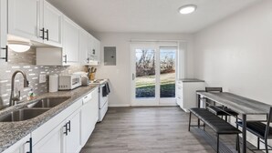 Een moderne keuken met witte kasten, een granieten aanrechtblad, een inbouwoven en een eethoek met een bankje en tafel.