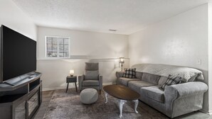 Een woonkamer met een flatscreentelevisie, een grijze bank, een salontafel, een grijze fauteuil, een klein bijzettafeltje en een lamp.