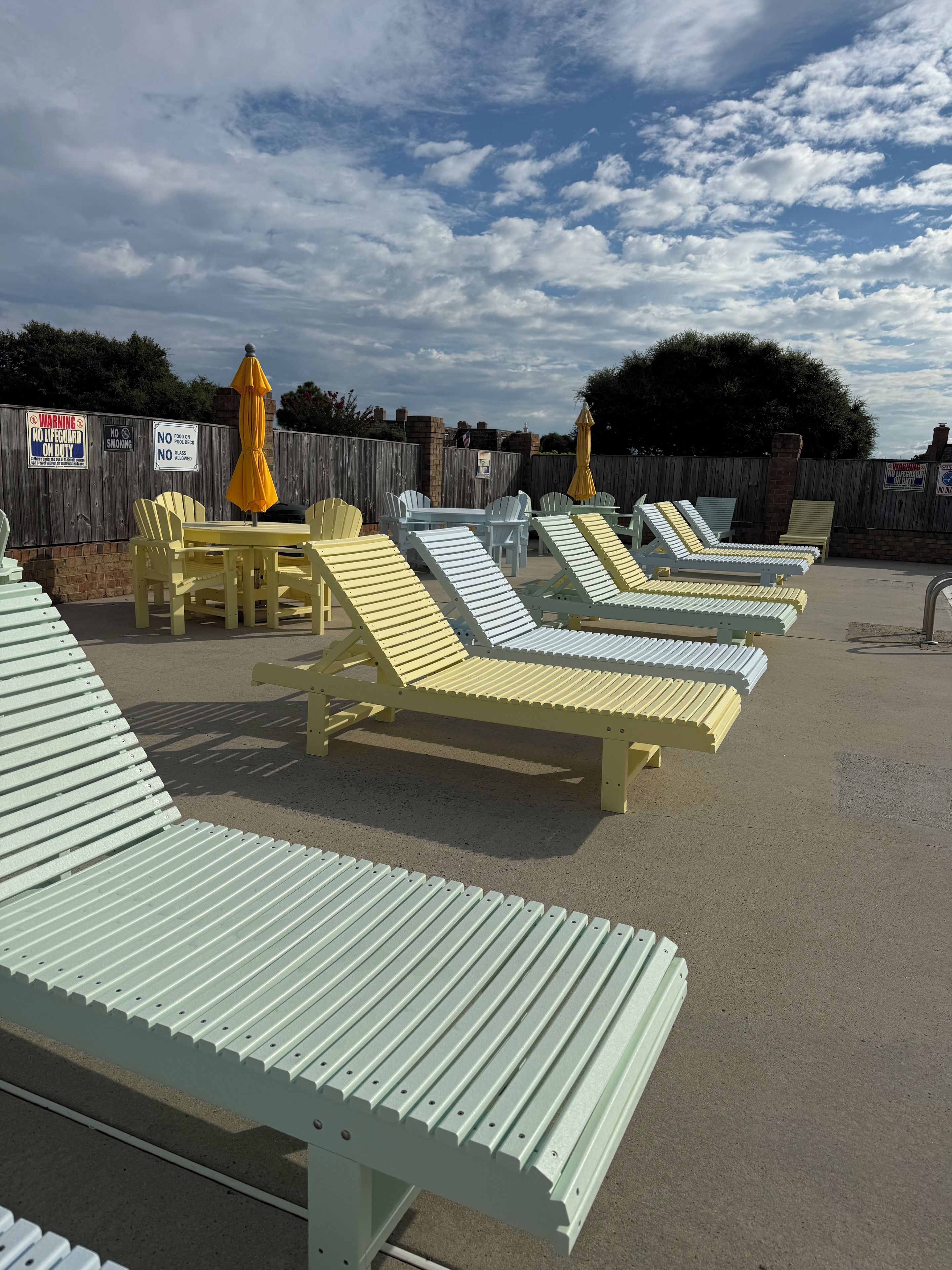 Des fauteuils et des tables d’extérieur sont disposés sur une surface en béton, avec une clôture en bois et des panneaux d’avertissement en arrière-plan.