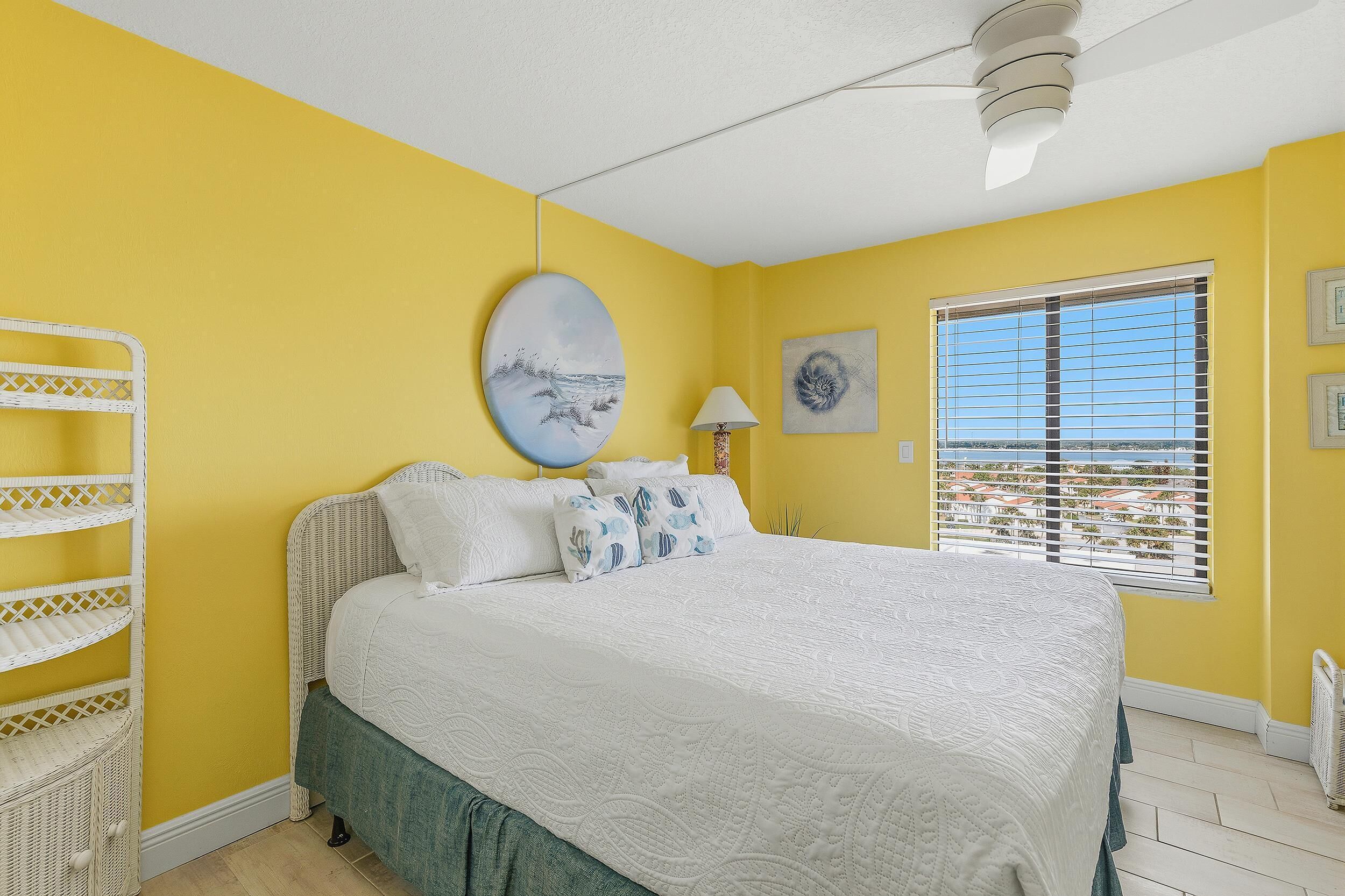 Un dormitorio con una cama, un ventilador de techo blanco, una ventana con persianas y una pared con un cuadro.