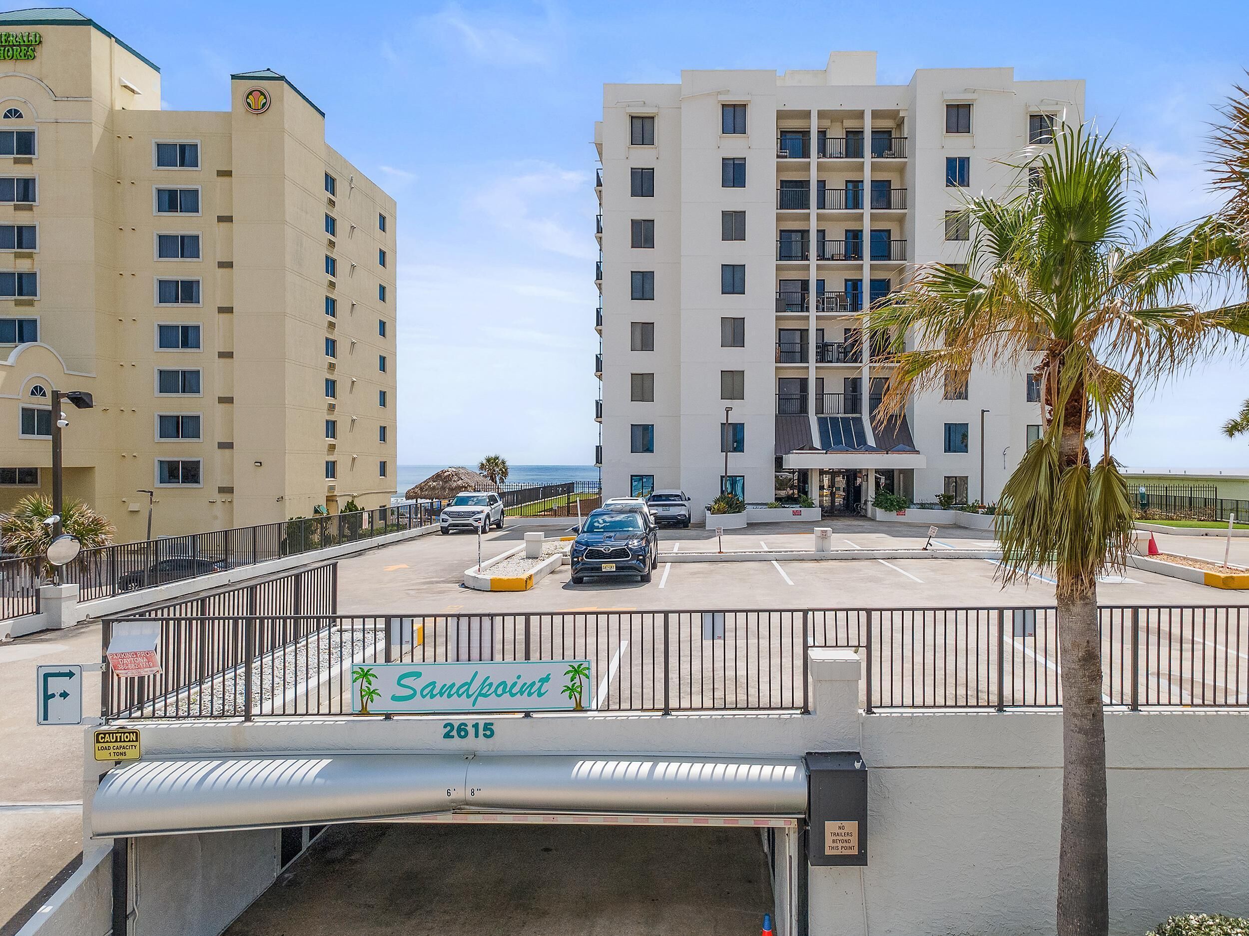 A hotel entrance with a sign reading 'Sandpoint 2615,' a parking area with cars, and a view of the ocean.
