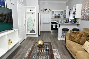 A modern living room with a television, a coffee table with a globe, a sofa, and a kitchen area with a refrigerator, microwave, and bar stools. - Cute as a Button, AC, Starlink, Wi-Fi, Hot Tub, Near Beaches, Bars & Restaurants (North Palmetto Point)