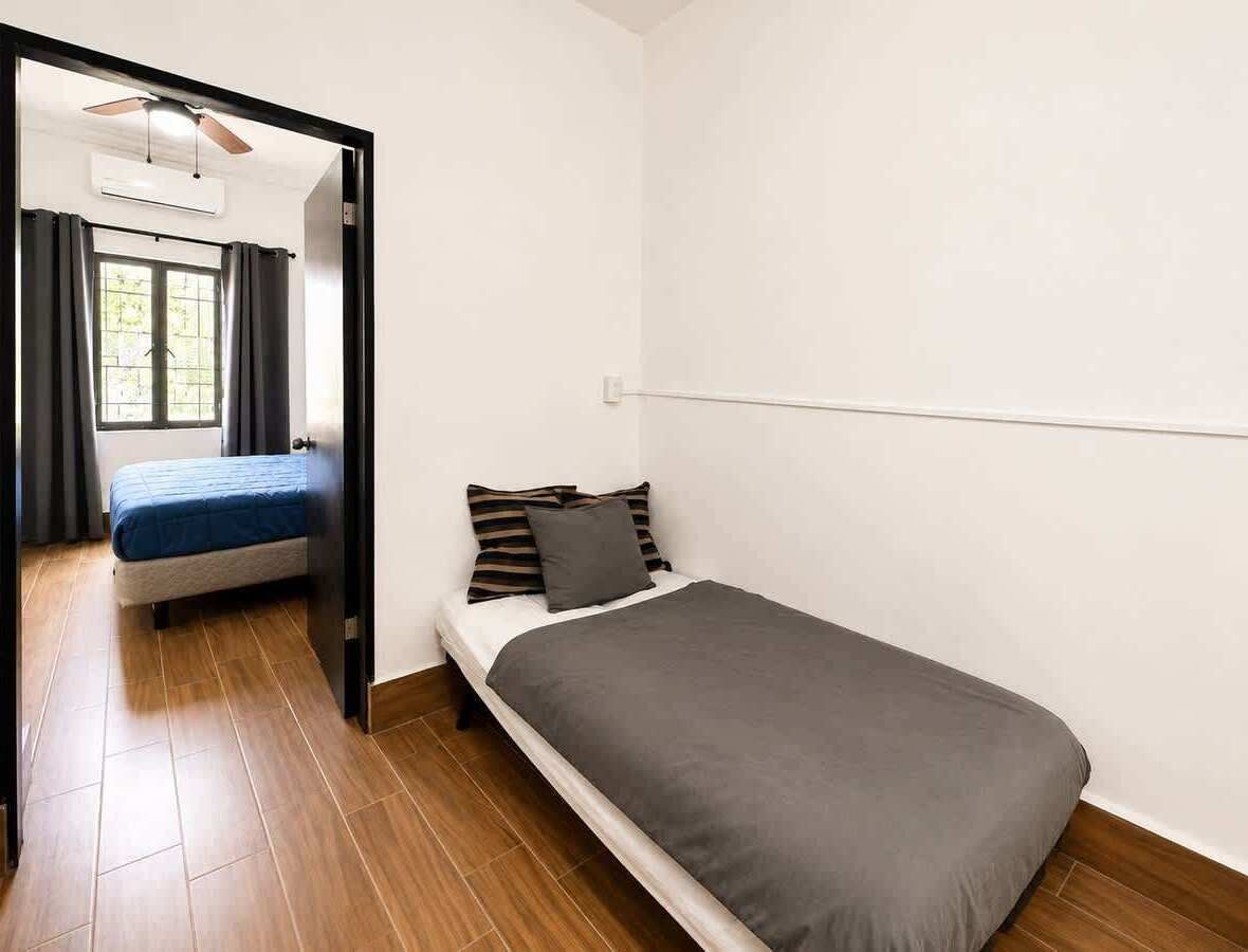 A small, single-bed room with a wooden floor, a window with curtains, and a ceiling fan.