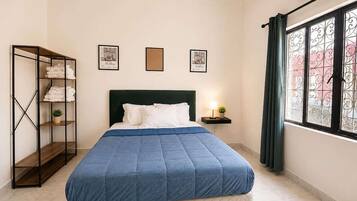 Ein Schlafzimmer mit einem Bett, einem Regal mit gefalteten Handtüchern, gerahmten Bildern an der Wand und einem Fenster mit Vorhängen.