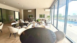 Un espacio interior moderno con mesas y sillas redondas, una piscina visible a través de amplios ventanales y una paleta de colores neutros.