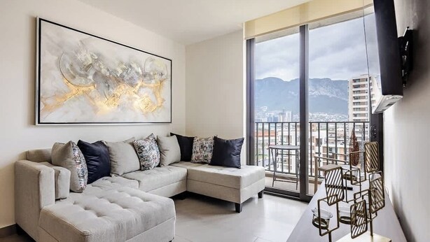 Una sala de estar moderna con un sofá modular, un cuadro abstracto de gran tamaño y vistas a un paisaje urbano a través de amplios ventanales.