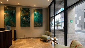 A lobby with a reception desk, three abstract paintings, a seating area with a sofa and armchairs, and large windows offering a view of the street. - Comfort Eden/Gym/Wi-fi/Parking/BBQ/ARENA (Monterrey)