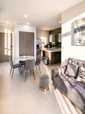 Una sala de estar moderna con un sofá gris, una mesa de comedor con sillas, una zona de cocina con refrigerador y estufa, y un cuadro abstracto grande en la pared.