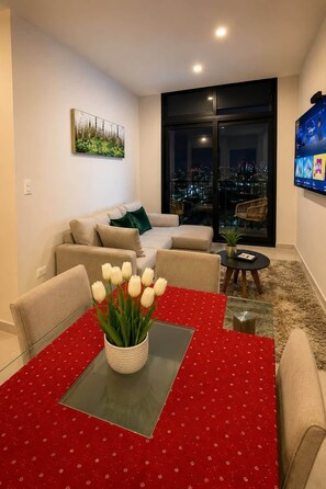 Ein modernes Wohnzimmer mit einer Couch, einem Esstisch mit roter Tischdecke und Blick auf die Stadt durch große Fenster.