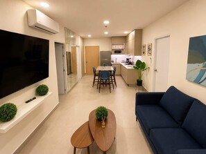 A modern living room with a flat-screen TV, a blue sofa, a wooden coffee table, and a dining area with a table and chairs.