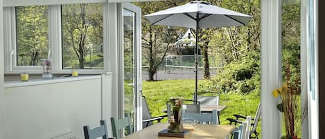 A conservatory with a table and chairs, a large umbrella, and a view of a green landscape.