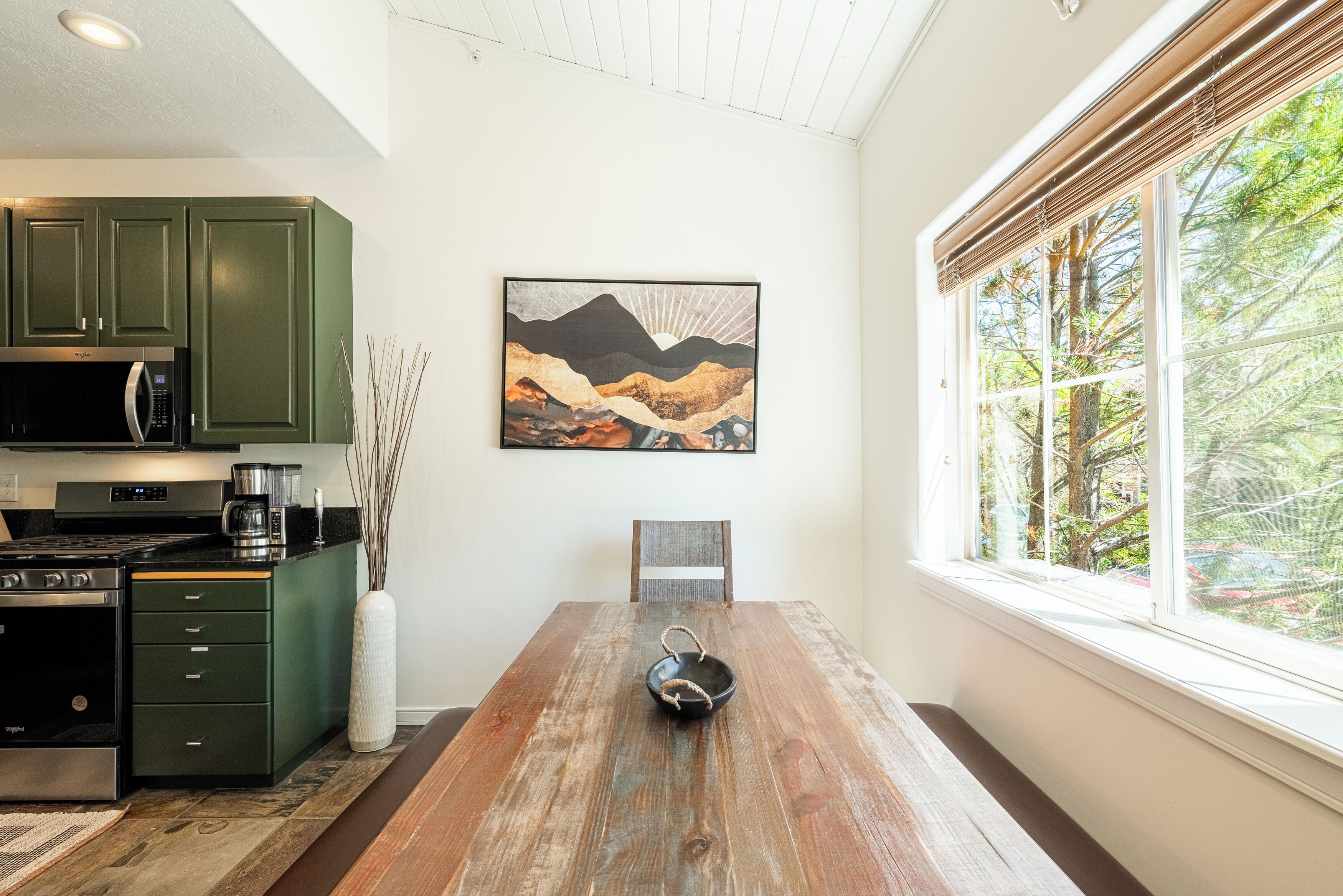 A dining area with a wooden table, a painting, and a view of trees through a window.