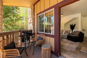 Eine überdachte Veranda mit Korbmöbeln, einem Grill und Blick in ein Wohnzimmer mit Sofa und Teppich.
