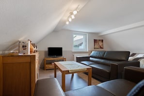 A loft-style room with a sloped ceiling, a wooden table, a leather sofa, a television on a stand, and a window providing natural light.