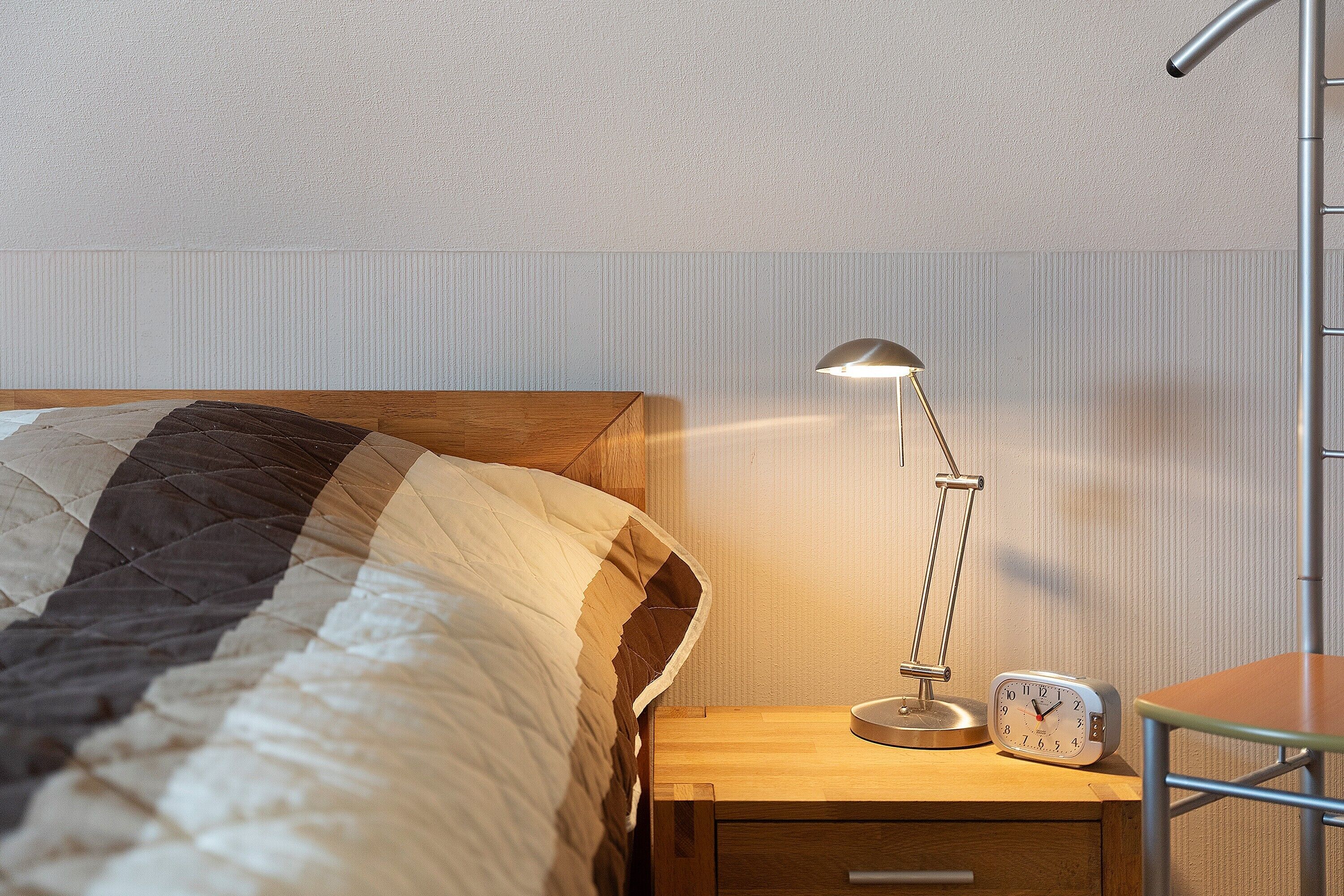 A neatly made bed with a striped blanket, a bedside table with a lamp and an alarm clock, and a chair.