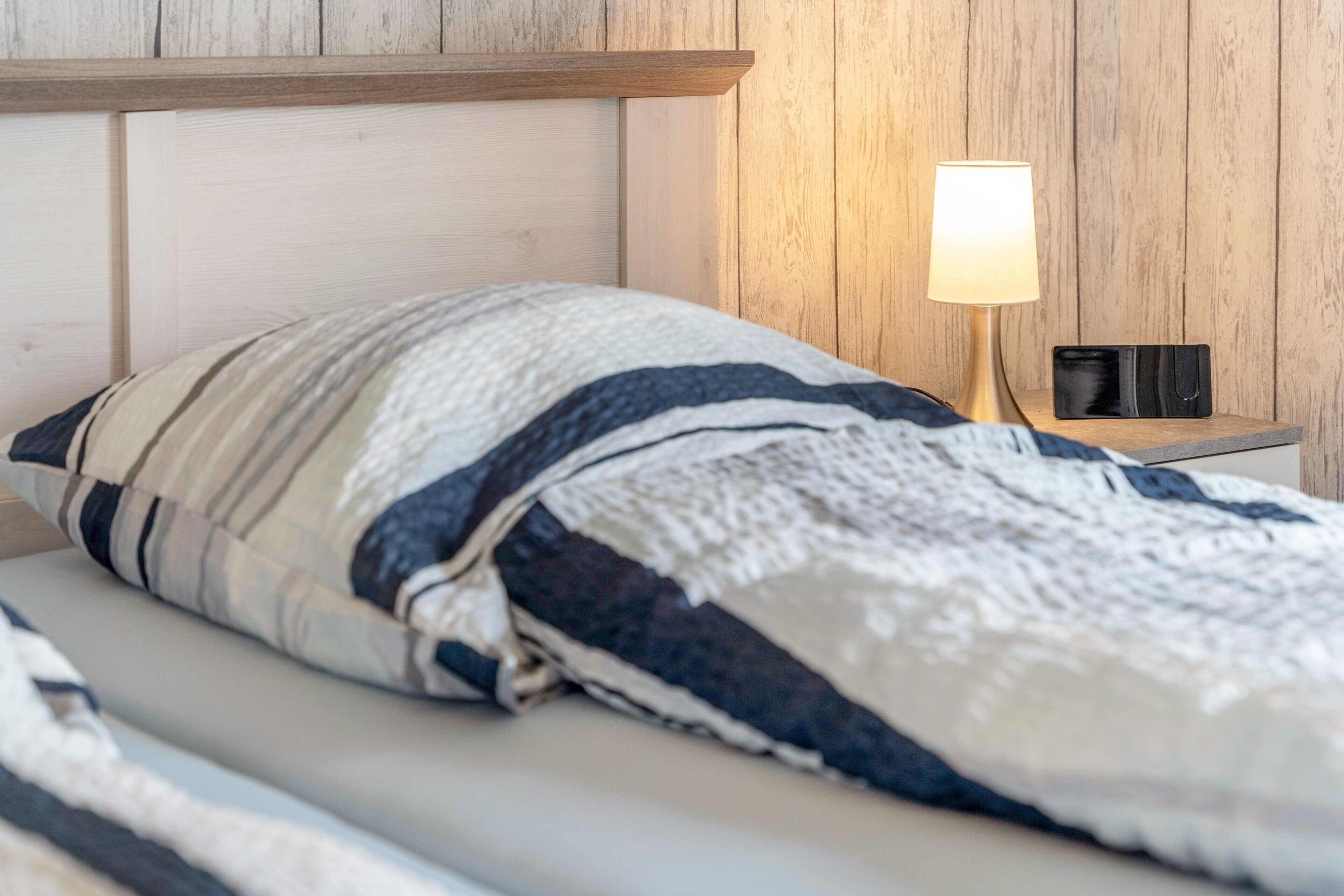 Ein Bett mit gestreiftem Kopfkissen und einer Decke, eine Nachttischlampe und ein kleines schwarzes Objekt auf dem Nachttisch.