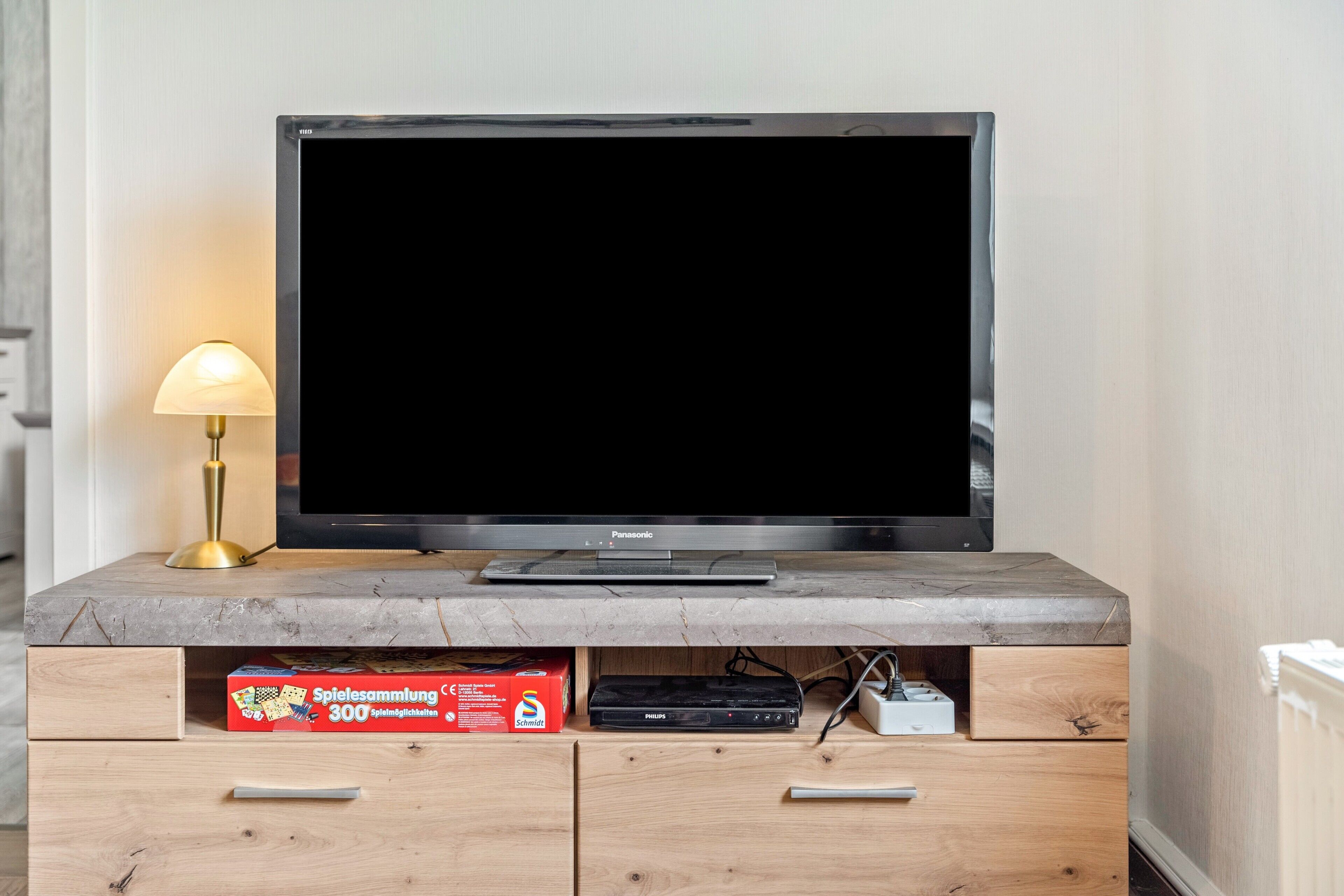 Ein Flachbildfernseher auf einem hölzernen TV-Schrank mit einer Lampe, einem Philips Gerät und einer Spielesammlung von Schottel.