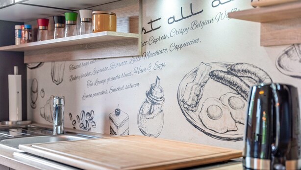 Una encimera de cocina con una tabla de cortar de madera, una cafetera y una pared decorada con ilustraciones y texto sobre comida.