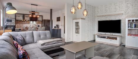 Ein modernes Wohnzimmer mit einem Ecksofa, einem Couchtisch und einem Flachbildfernseher. Es gibt eine Küchenzeile mit einer Mikrowelle und einer Kaffeemaschine.