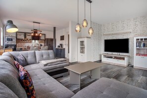 A modern living room with a sectional sofa, a coffee table, and a flat-screen TV. There is a kitchen area with a microwave and a coffee machine.