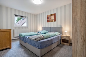 A bedroom with a bed, bedside table, lamp, window, radiator, and striped wallpaper.