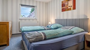 Un dormitorio con cama, ventana, radiador, cómoda de madera y una obra de arte en la pared.