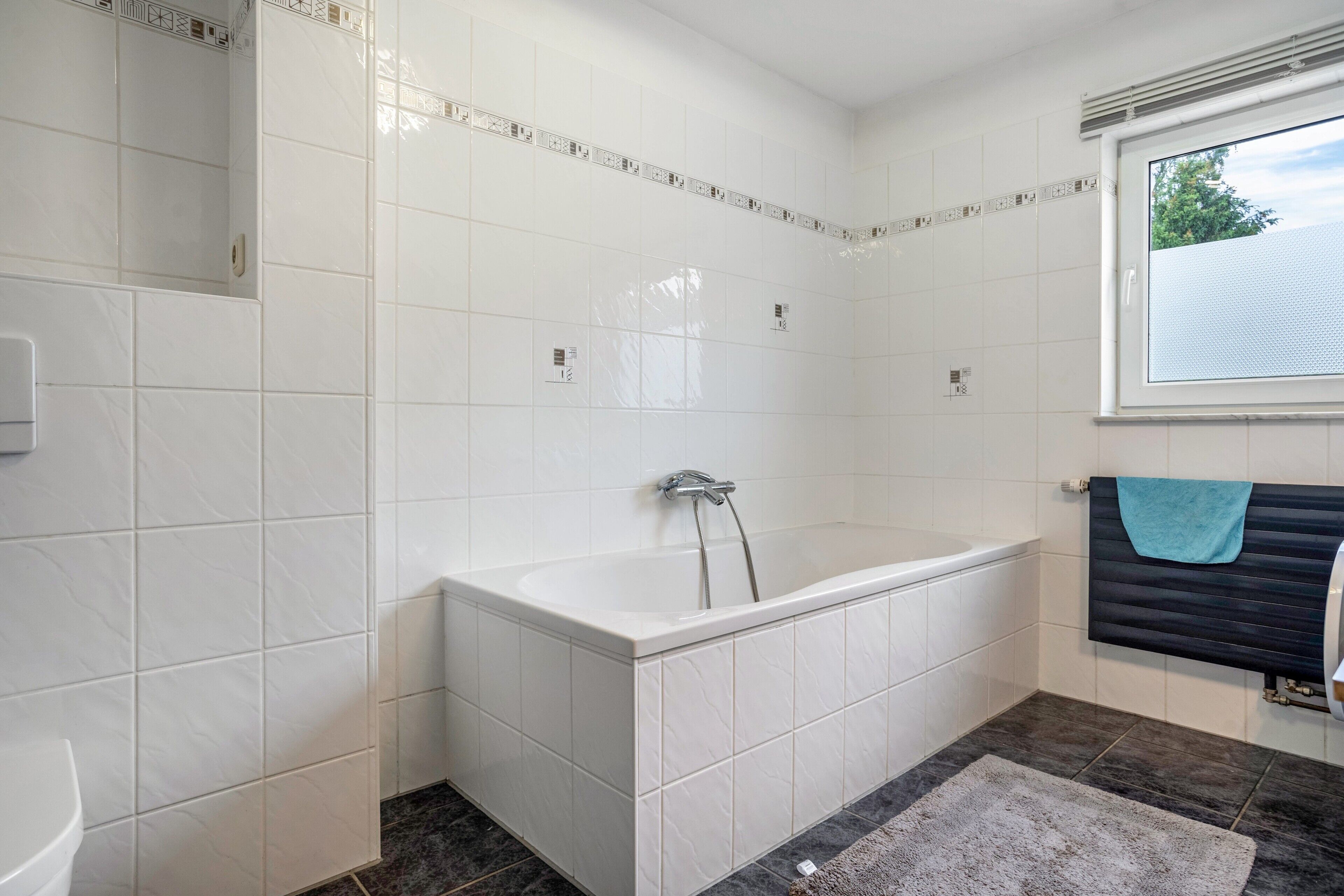 A bathroom with white tiles, a bathtub, a window, and a towel rack.