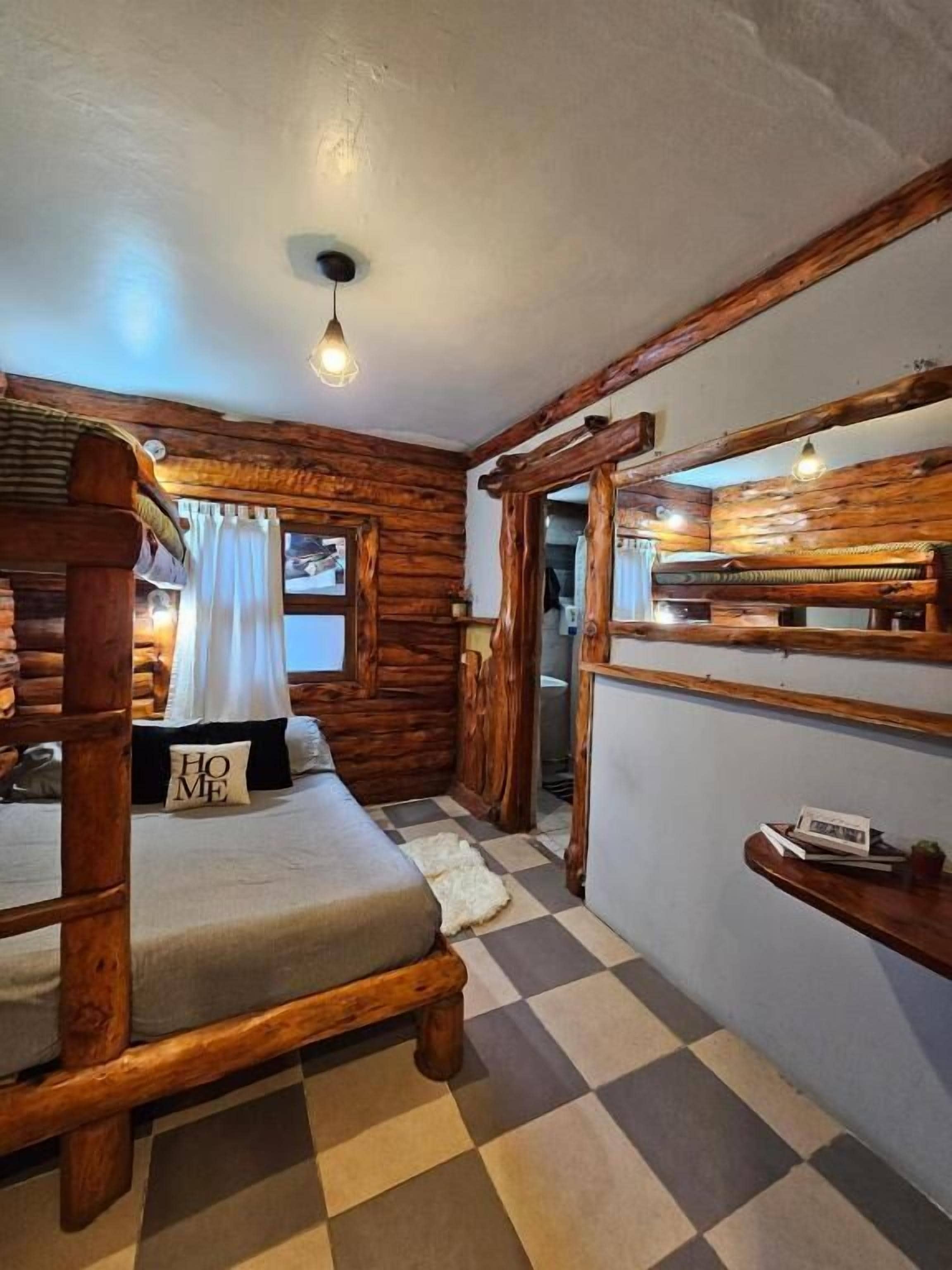 A log cabin-style room with a bed, a checkered floor, wooden walls, and a small shelf.